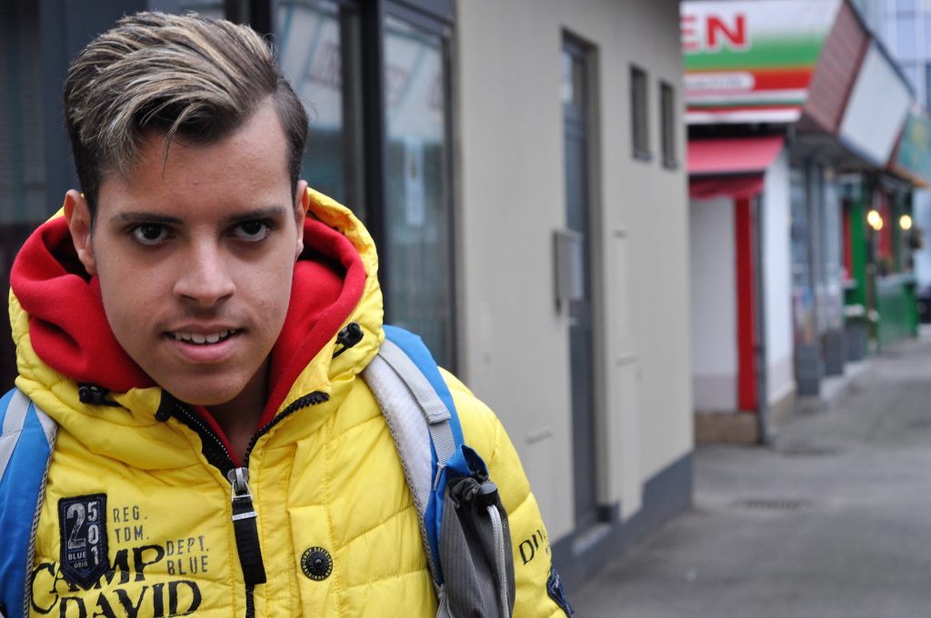 Portraitfoto von Soufiene. Er trägt eine gelbe Jacke, hat kurze Haare, schaut in die Kamera und lächelt leicht. Am Rücken seine Schultasche.