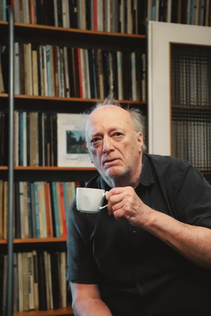 Josef (Mann mit lichtem weissen Haar) sitzt mit einer weissen Tasse Kaffee in der Hand vor einen regal mit Schallplatten