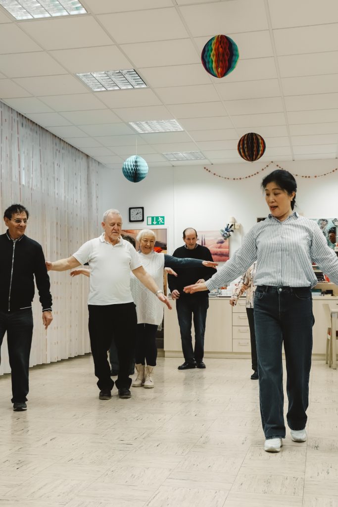 Auf dem Bild ist die Tanzlehrerin im Vordergrund zu sehen. Sie trägt Jeans und ein blaues Hemd. Im Hintergrund einige Senior*innen. Sie üben gerade Schritte.