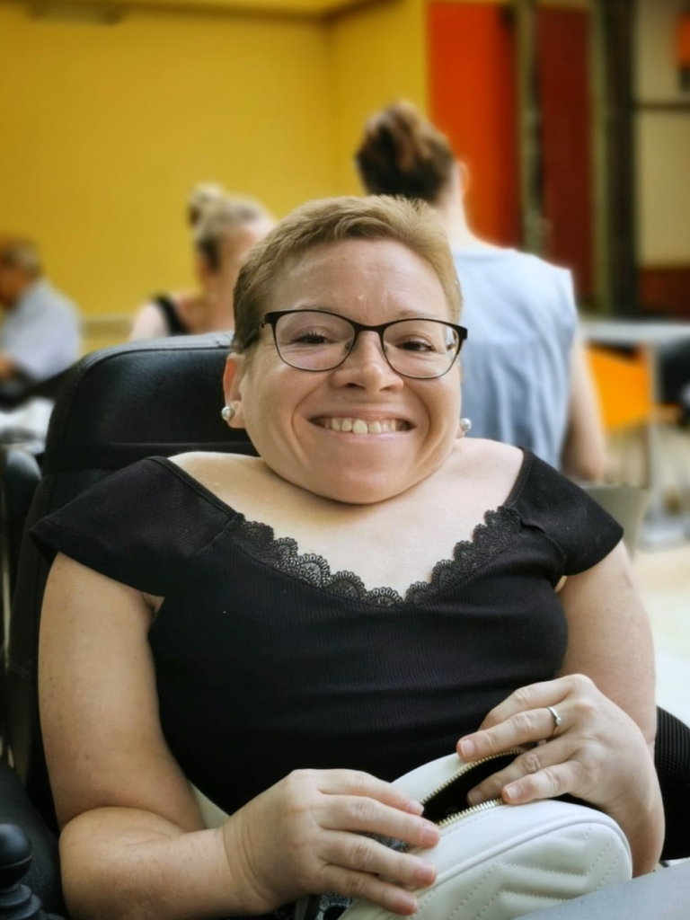 Porträt von Evelyn. Eine Frau mit kurzen Haaren und schwarzer Brille. Sie nutzt einen Rollstuhl.