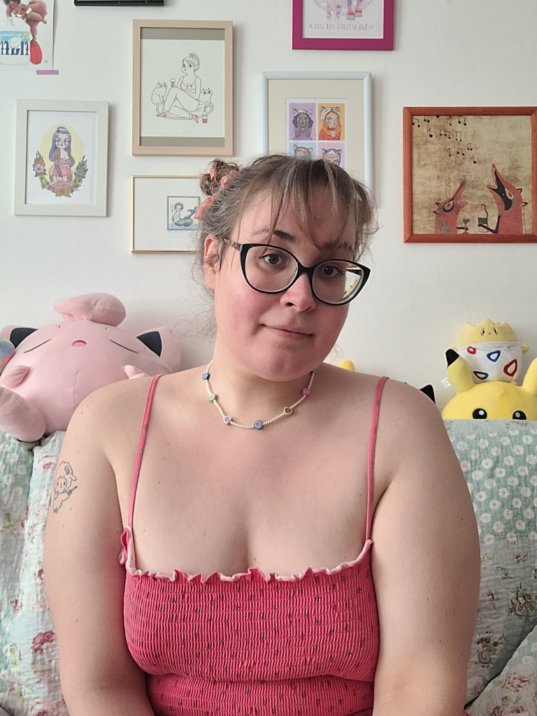 Porträt von Lena. Sie träge ein rotes Top mit Spagetthiträgern, eine schwarze Brille und einen Pony. Sie sitzt auf einer Couch in ihrem Zimmer. Hinter ihr bunte Bilderrahmen und Kuscheltiere.