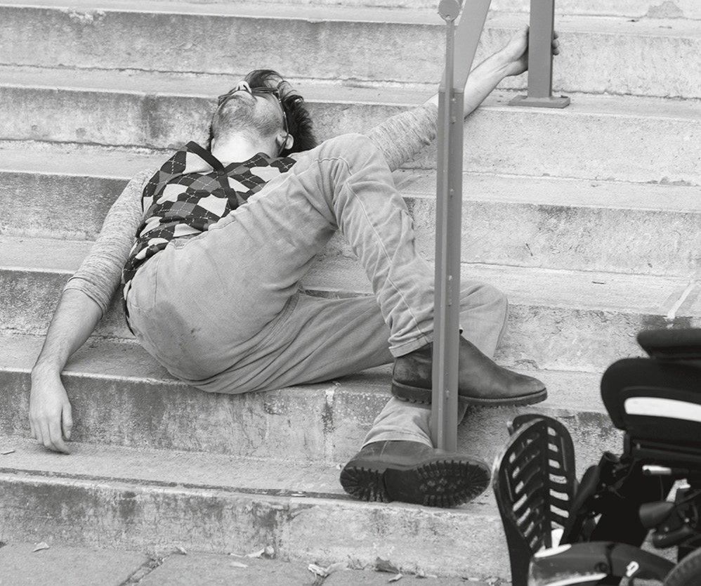 Ein Mann liegt auf den Stufen einer Treppe. Vorne im Bild ist ein Teil seines Rollstuhls zu sehen.