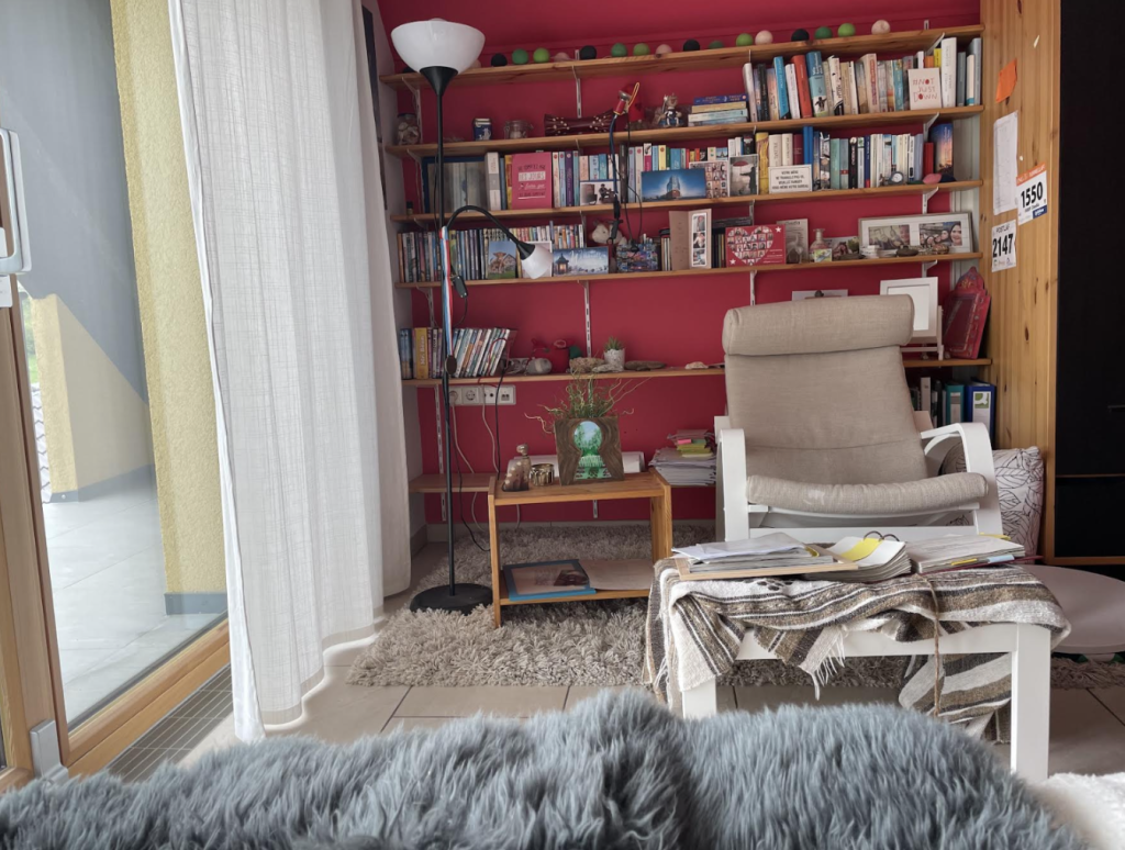 Blick vom Bett auf ein Bücherregal vor einer roten Wand. Ein Sessel und ein Vorhang zum Fenster