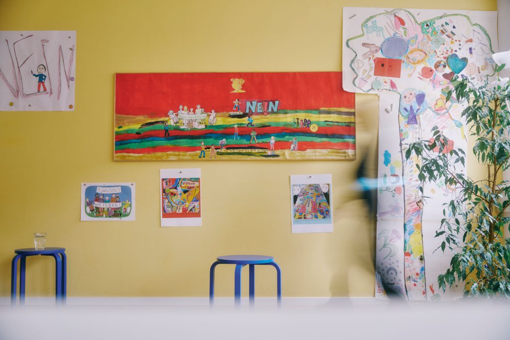 Das Foto zeigt eine gelbe Wand, an der bunte Plakate mit dem Titel "Nein" hängen. Davor stehen blaue Stühle und rechts eine Zimmerpflanze.