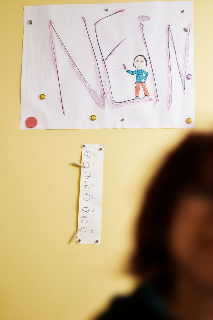 Links unten sieht man verschwommenen Umrisse eines Gesichts. Im Hintergrund hängt ein Plakat mit dem Titel "Nein". Im Hintergrund ist eine gelben Wand.
