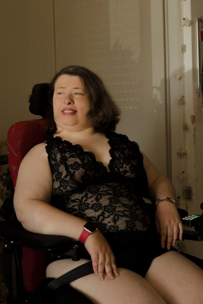 Leonie Schüler sitzt in ihrem Rollstuhl. Sie schaut nach links. Sie trägt einen schwarzen Spitzen-Body und pinken Lippenstift.