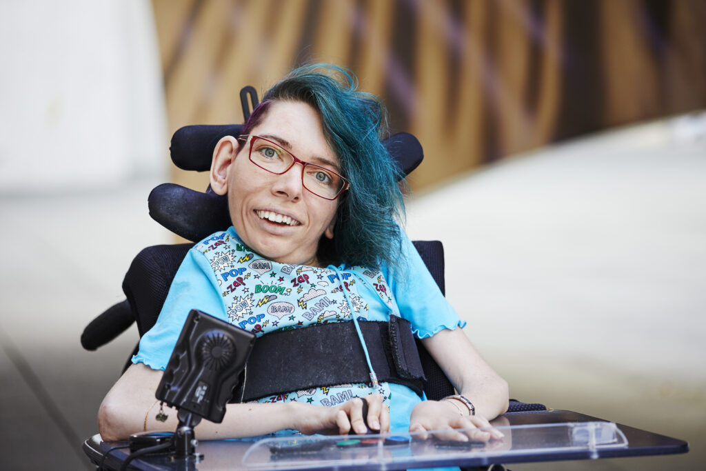 Melanie Eilert schaut mit freundlichem Blick in die Kamera. Sie hat blaue Haare, trägt eine Brille und sitzt in einem Rollstuhl.