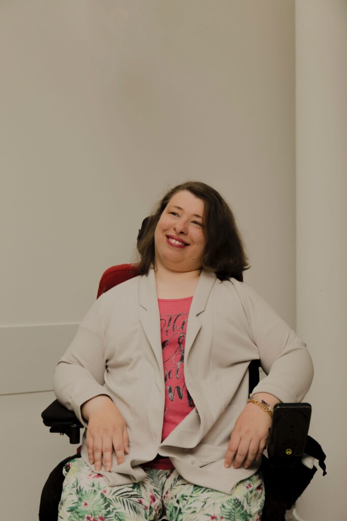 Leonie Schüler schaut in lachend in die Kamera. Sie trägt einen beigen Blazer und pinken Lippenstift. Sie nutzt einen Rollstuhl.