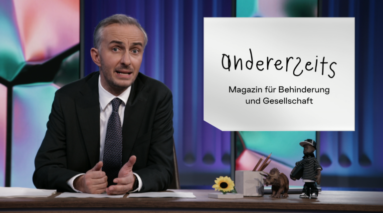 Das Bild zeigt Jan Böhmermann, einen bekannten Moderator, der hinter einem Tisch sitzt. Er trägt einen schwarzen Anzug, ein weißes Hemd und eine gestreifte Krawatte. Rechts neben ihm steht ein Schild mit der Aufschrift „andererseits – Magazin für Behinderung und Gesellschaft“. Auf dem Tisch liegen Papiere, ein kleiner Dinosaurier, ein kleines Männchen in Baseballkleidung, eine gelbe Blume und eine Box mit Stiften. Der Hintergrund ist bunt und modern gestaltet.