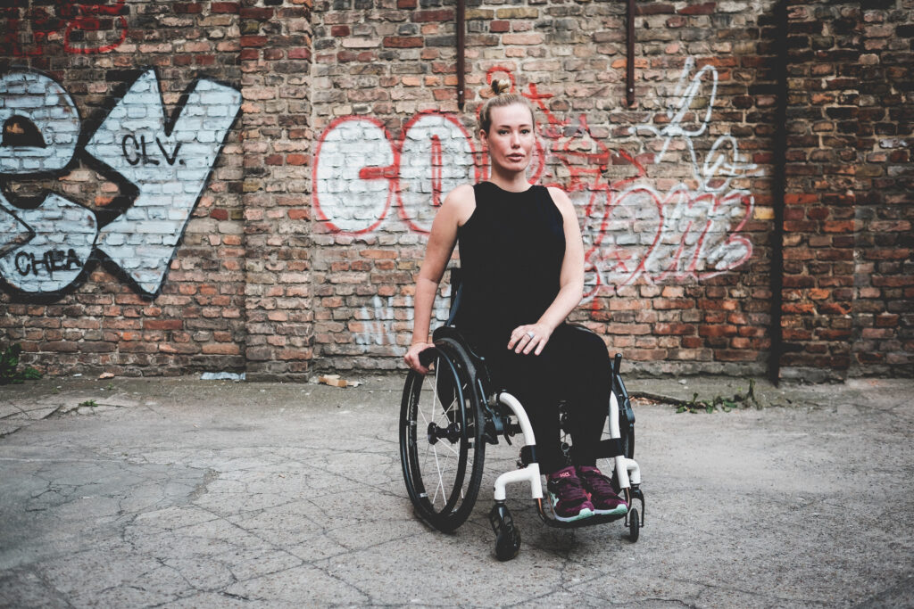 Eine blonde Frau sitzt in einem Aktiv-Rollstuhl. Sie ist komplett schwarz gekleidet. Ihre Haare trägt sie zusammengebunden zu einem hohen Knoten. Im Hintergrund ist eine Ziegelmauer mit Graffitti zu sehen.