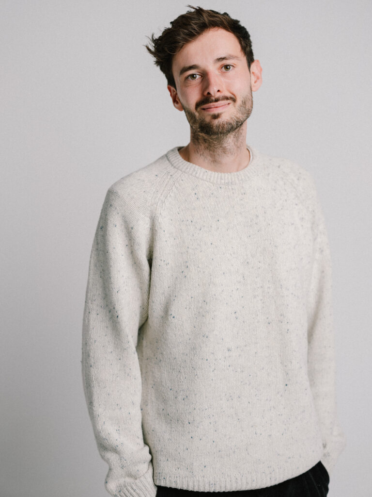 Ein junger Mann in weißem Pullover schaut in die Kamera.