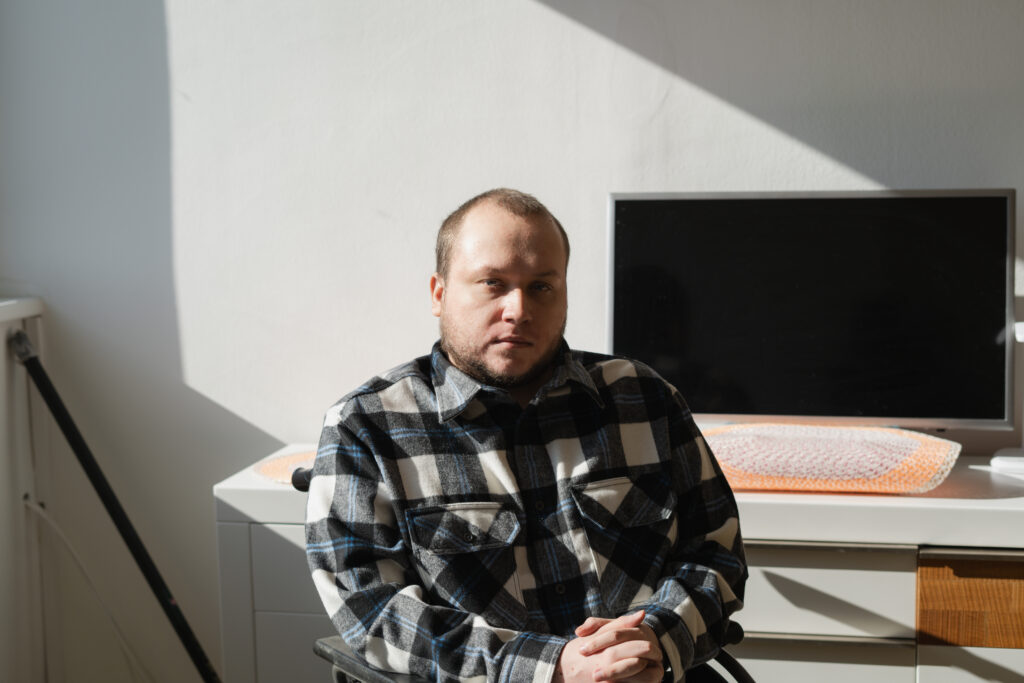 Anton schaut mit ernstem Blick in die Kamera. Er hat kurze, braune Haare und trägt ein Karo-Hemd. Hinter ihm steht ein Computer-Bildschirm auf einem Schreibtisch.