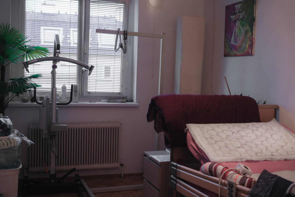 Ein Zimmer mit einem Pflegebett. Das Bett hat eine dunkelrote Decke und ein weißes Kissen. Neben dem Bett steht ein Lifter für Menschen mit Mobilitätseinschränkungen. Vor dem Fenster stehen einige Pflanzen.