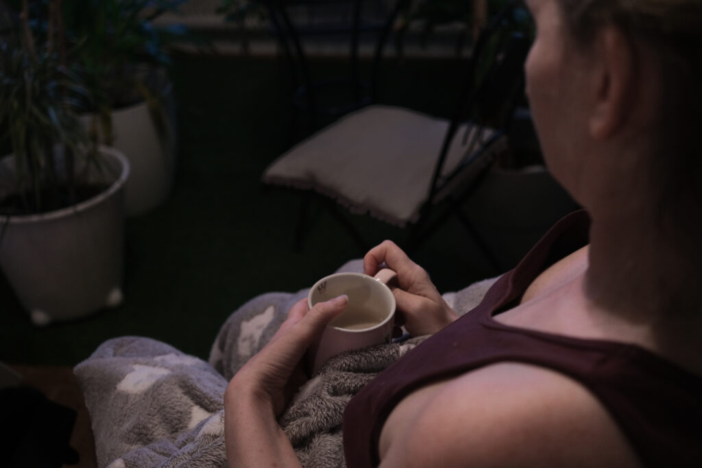 Claudia Turetscheck sitzt mit einer grauen Decke auf einem Stuhl und hält eine Tasse in den Händen. Sie trägt ein dunkles Oberteil. Im Hintergrund stehen Pflanzen. Die Beleuchtung ist warm und gedimmt.
