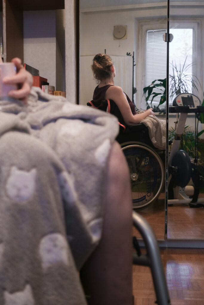 Claudia Turetscheck sitzt in einem Rollstuhl vor einem Spiegel. Sie schaut aus dem Fenster. Im Vordergrund sieht man die Reflexion ihres Spiegelbildes. Ihre Beine sind mit einer grauen Decke umwickelt.