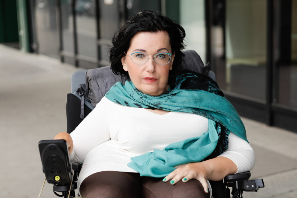 Dorothea Brozek sitzt in einem elektrischen Rollstuhl. Sie hat dunkle Haaren und trägt eine Brille. Sie trägt ein weißes Oberteil und einen türkisfarbenen Schal. Ihre Nägel sind passend lackiert. Sie schaut direkt in die Kamera. Im Hintergrund sind Glasfenster und ein Gehweg.