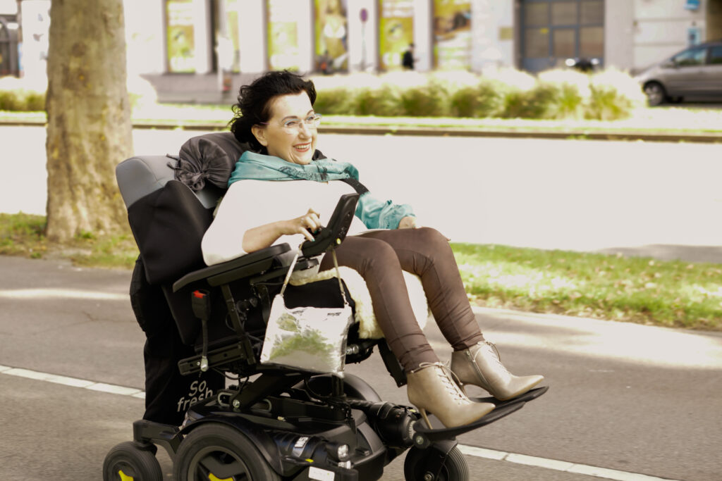 Das Bild zeigt Dorothea Brozek, sie sitzt in einem elektrischen Rollstuhl und fährt auf dem Gehsteig. Sie lacht freundlich. Im Hintergrund sind mehrere Bäume und eine Straße.