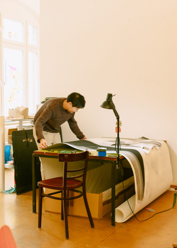 Juewen Zhang steht an einem Tisch in seinem Atelier. Er betrachtet große Drucke oder Pläne und arbeitet konzentriert. Der Raum ist hell.