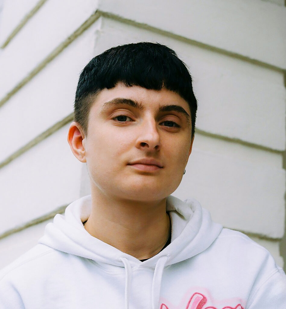 Eine Person mit kurzen schwarzen Haaren schaut mit ernstem Blick in die Kamera. Sie trägt einen weißen Pullover. Die Person steht vor einem weißen Gebäude.