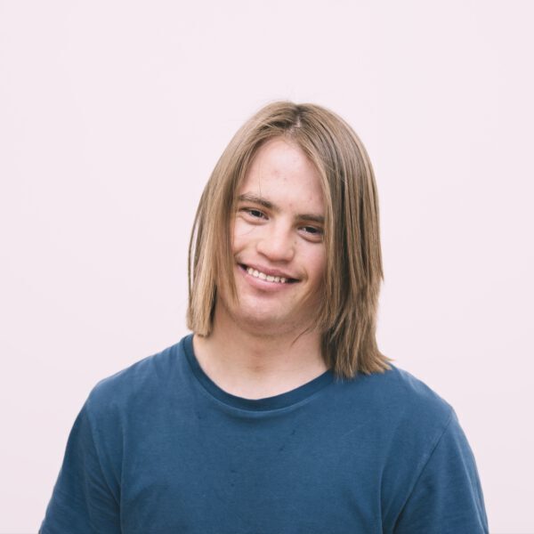 TIll Krummwiede lächelt in die Kamera. Er hat schulterlange dunkelblonde Haare. Der Hintergrund ist rosa.