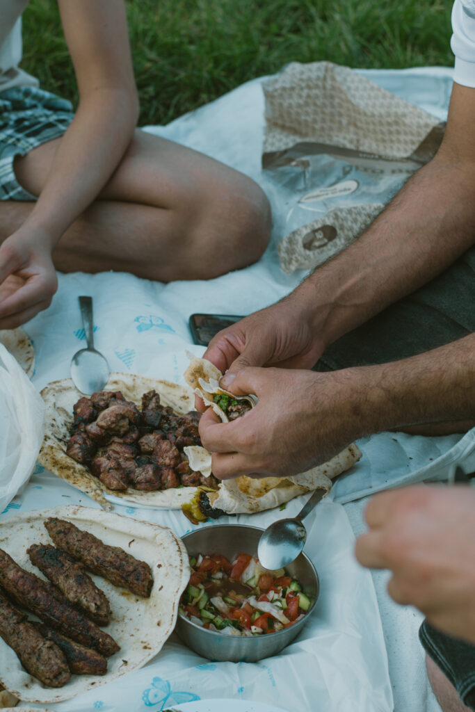Das Foto zeigt ein Picknick. Auf einer weißen Decke sitzen Personen. In der Hand halten sie Fladenbrot. Auf der Decke ist ein Salat und Lammspieße.